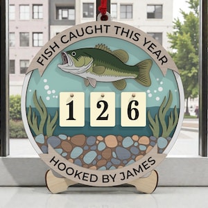 May include: A wooden fishing-themed ornament with the text "FISH CAUGHT THIS YEAR" and "HOOKED BY JAMES". It features a green fish illustration, a counter showing "126", and aquatic elements.