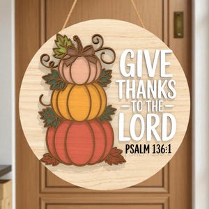May include: A round wooden sign with a stacked pumpkin design in shades of pink, orange, and red. The sign reads "GIVE THANKS TO THE LORD" with "PSALM 136:1" below. A jute rope is attached for hanging. The sign is displayed on a wooden door.