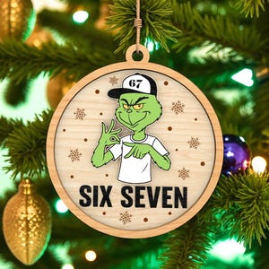 May include: Wooden Christmas ornament featuring a green cartoon character wearing a baseball cap and white shirt. The ornament has the text "SIX SEVEN" and snowflake accents. The ornament is hanging on a Christmas tree.