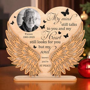 May include: A wooden memorial plaque shaped like a heart, with a black and white photo of a man. The plaque has laser-cut angel wings and the text "My mind still talks to you and my Heart still looks for you but my soul knows you're AT PEACE."