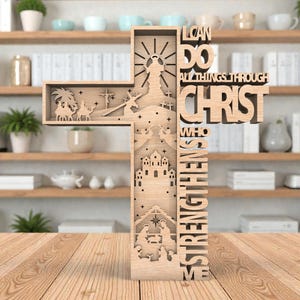 May include: A wooden cross with intricate cut-out designs depicting religious scenes and the text "I CAN DO ALL THINGS THROUGH CHRIST WHO STRENGTHENS ME." The cross is made of light-colored wood and stands on a wooden surface.