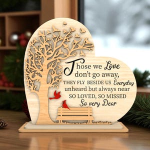 May include: A wooden heart-shaped memorial plaque with a tree and birds cut-out design. The plaque features the text "Those we Love don't go away..." and two red bird accents. The base is rectangular.