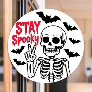 May include: Round white Halloween sign with a skeleton giving a peace sign. The sign has the words "STAY Spooky" in red, with black bat silhouettes around the skeleton.