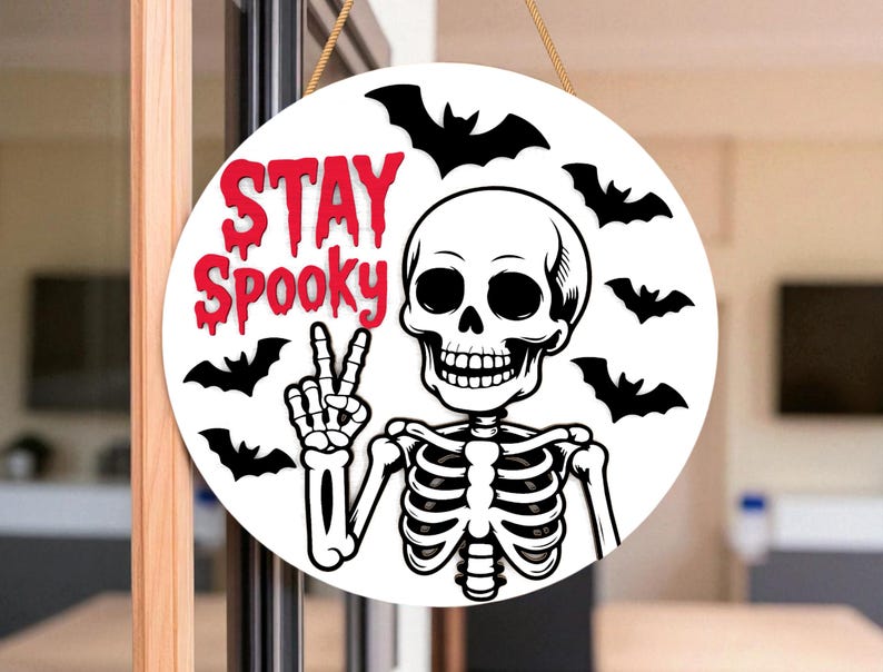 May include: Round white Halloween sign with a skeleton giving a peace sign. The sign has the words "STAY Spooky" in red, with black bat silhouettes around the skeleton.