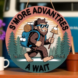 May include: Round wooden sign with the text "S'more Advantres A Wait". The sign features a cartoon Bigfoot character wearing sunglasses and carrying a backpack, holding a stick with marshmallows. The background includes trees and a sunset.