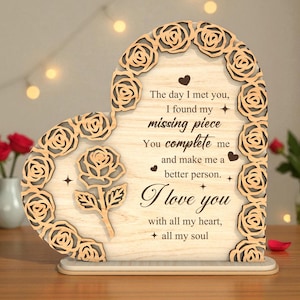 Puede incluir: Adorno de madera en forma de corazón con un diseño de rosas cortadas con láser y un borde de rosas. El texto "El día que te conocí, encontré mi pieza que faltaba... Te amo" está grabado.