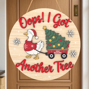 Peut inclure: Panneau rond en bois avec l'inscription "Oops! I Got Another Tree" en rouge. Un dessin d'oie portant un bonnet de Père Noël et un pull tire une charrette rouge avec un sapin de Noël. Des flocons de neige ornent le design.