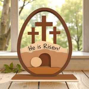 Op de afbeelding: Houten paasei-decoratie met drie kruisen, een graf en de tekst "He is Risen!" in bruin, beige en crème. De decoratie staat op een houten voet, tegen een raam met een vage groene achtergrond.