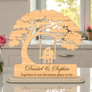 May include: A wooden tree-shaped decorative piece with a swing featuring two figures. The base reads "Daniel & Sophia" and "Together is our favourite place to be". The piece is light brown and sits on a cream-colored base.