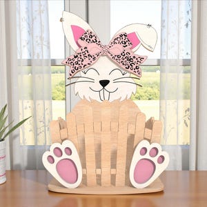 Puede incluir: Cesta de Pascua de madera con forma de conejito con un lazo rosa y estampado de leopardo. El conejito tiene orejas y patas blancas con detalles rosas. La cesta es de madera marrón claro.