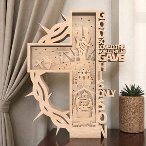 Puede incluir: Decoración religiosa en forma de cruz de madera con intrincados diseños recortados. Presenta una corona de espinas, una escena de crucifixión y el texto "GOD SO LOVED THE WORLD THAT HE GAVE HIS ONLY SON". Una planta suculenta decorativa está cerca.