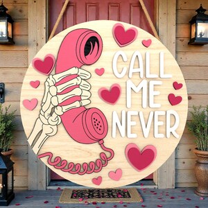May include: A round wooden sign with a pink telephone held by a skeleton hand, surrounded by pink hearts. The words "CALL ME NEVER" are in white. The sign hangs from a rope.