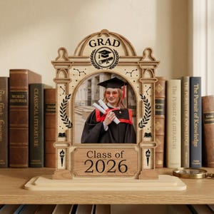 May include: Wooden graduation photo frame with a picture of a graduate in a cap and gown. The frame is engraved with the word "GRAD", a diploma, and "Class of 2026". The frame is surrounded by books.