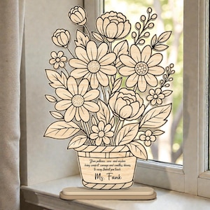 Puede incluir: Arreglo floral de madera con un jarrón, con margaritas, rosas y otras flores. El jarrón tiene un mensaje personalizado y el nombre "Ms. Fank". El arreglo se asienta sobre una base de madera, ideal para un regalo de profesor.