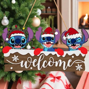 Puede incluir: Un letrero navideño de madera con tres personajes de dibujos animados con gorros de Papá Noel y guantes rojos. El letrero dice "welcome" en escritura blanca, con copos de nieve. Los personajes son azules con orejas grandes y boca abierta.