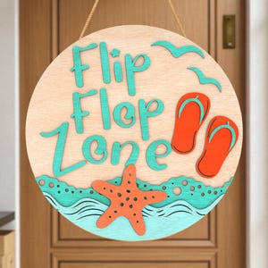 Puede incluir: Cartel de madera redondo con las palabras "Flip Flop Zone" en turquesa. El cartel presenta chanclas naranjas, una estrella de mar y gaviotas. El cartel está colgado de una cuerda.
