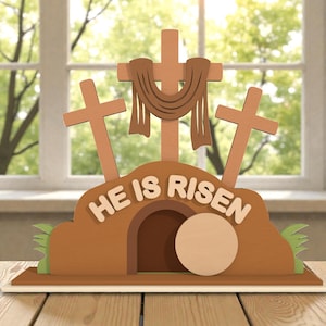 May include: A wooden Easter decoration featuring three crosses, a tomb, and a stone. The words "HE IS RISEN" are displayed on the tomb. The decoration is brown with tan and green accents, set against a window background.