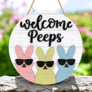May include: Round wooden sign with the words "welcome Peeps" in black script. The sign features three pastel-colored bunny silhouettes wearing sunglasses. A brown rope is attached for hanging.