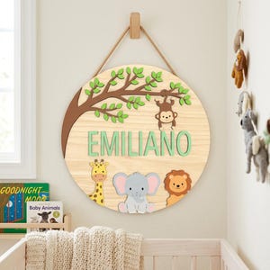 May include: A circular wooden sign with a jungle theme. It features a tree branch, a monkey, a giraffe, an elephant, and a lion. The name "EMILIANO" is displayed in green letters. The sign is suspended on a wall.