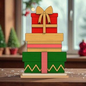 Op de afbeelding: Een houten kerstdecoratie met een stapel van vier cadeau dozen. De bovenste doos is rood met een gouden strik, gevolgd door een lichtbruine doos, een roze doos en een groene doos met een gele zigzag ontwerp. De decoratie staat op een houten voet.