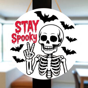 May include: A round, white Halloween sign with the words "STAY Spooky" in red dripping font. A black and white skeleton is giving the peace sign, surrounded by black bat silhouettes. The sign is hanging by a natural rope.