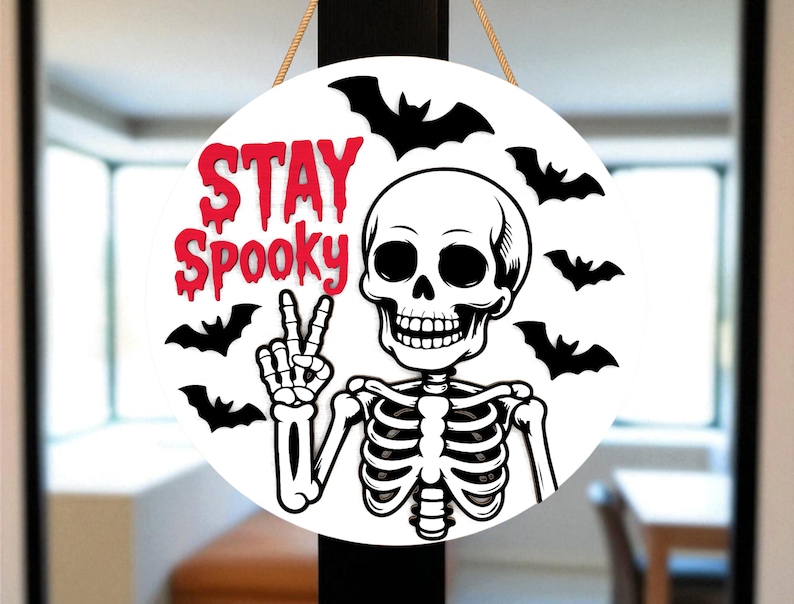 May include: A round, white Halloween sign with the words "STAY Spooky" in red dripping font. A black and white skeleton is giving the peace sign, surrounded by black bat silhouettes. The sign is hanging by a natural rope.