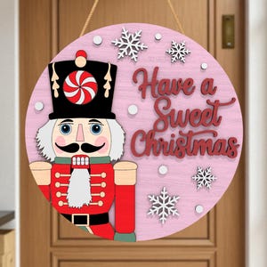 May include: A round, pink wooden Christmas sign featuring a nutcracker soldier and the phrase "Have a Sweet Christmas" in red script. The sign includes white snowflakes and a candy design on the nutcracker's hat. A gold string is attached for hanging.