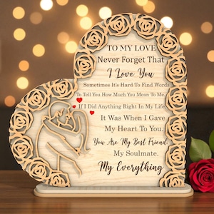 May include: Wooden heart-shaped plaque with a floral border and a loving message. The plaque features a cutout design of a couple embracing and the words "To My Love, I Love You, My Everything." Two red roses are in the foreground.