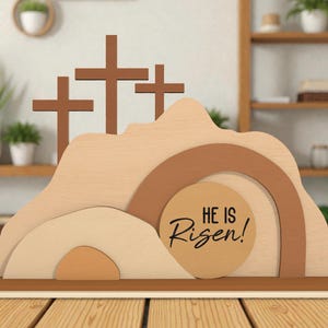 Puede incluir: Decoración de Pascua de madera con tres cruces marrones sobre un paisaje en capas. El diseño incluye un arco beige y un elemento circular con el texto "HE IS Risen!" en escritura negra. La base es marrón.