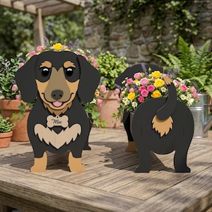 Op de afbeelding: Houten teckel hondenplanter met een zwart en bruin lichaam, met een bloemstuk. De hond heeft een label met de naam "Max". De planter staat op een houten tafel, met andere planten op de achtergrond.