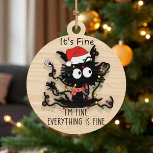 May include: Round wooden Christmas ornament with a black cat wearing a Santa hat and a scarf, tangled in fairy lights. The text "It's Fine, I'm Fine, Everything is Fine" is printed on the ornament. The ornament is hanging on a Christmas tree.