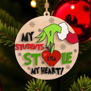 May include: Wooden Christmas ornament with the Grinch's hand reaching for a red apple with the word "LISA" on it. The ornament reads "MY STUDENTS STOLE MY HEART!" in black, red, and green lettering. Snowflakes are also featured.