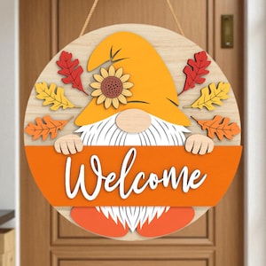 Könnte beinhalten: Runder Holz-Willkommensschild mit einem Zwerg mit orangefarbener Mütze und Bart, der ein orangefarbenes Banner mit dem Wort "Welcome" in weißer Schrift hält. Das Schild ist mit Herbstblättern und einer Sonnenblume verziert.