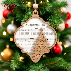 May include: Wooden Christmas tree ornament with the text "O Christmas Tree" and musical notes. The ornament features a cutout Christmas tree design and is hanging on a decorated Christmas tree with red and gold ornaments.