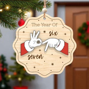 May include: Wooden Christmas ornament with a scalloped edge. The ornament features Santa's hands signing the numbers six and seven. The text "The Year Of * six * seven" is printed on the ornament.