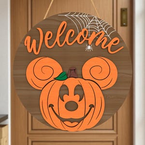 May include: Round wooden Halloween welcome sign featuring an orange pumpkin with Mickey Mouse ears and a smiling face. The word "Welcome" is in orange script above the pumpkin, with a spider and web design.