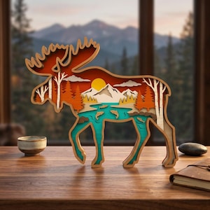 May include: A multi-layered wooden moose silhouette with a scenic landscape design. The artwork features a mountain range, trees, a lake with fish, and a setting sun, all within the moose's form. The color palette includes shades of orange, brown, white, and teal.