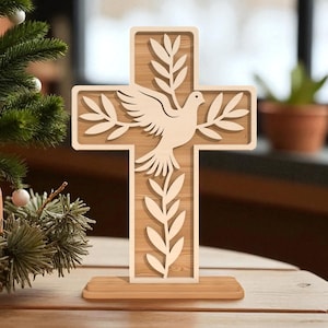 Può includere: Una croce di legno con un design a colomba e ramoscelli d'ulivo. La croce è marrone chiaro con un bordo più chiaro e una base di legno. La colomba è al centro della croce e i ramoscelli d'ulivo sono su entrambi i lati. La croce è su un tavolo di legno.