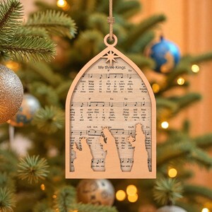 May include: Wooden Christmas ornament with a cutout design of the three wise men. The ornament is shaped like an arched window and features sheet music with the text "We three Kings". The ornament hangs from a natural twine string.