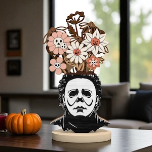 Pode incluir: Decoração de Halloween em madeira com um busto estilizado de Michael Myers e um buquê de flores. As flores são em tons de rosa, branco e marrom, algumas com detalhes de caveiras e abóboras. O busto está sobre uma base redonda e clara.