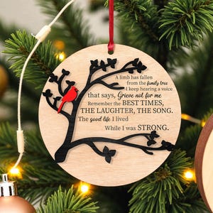 Könnte beinhalten: Runder Weihnachtsbaumschmuck aus hellem Holz mit einem schwarzen Baumdesign und einem roten Kardinal. Der Anhänger hat ein rotes Band zum Aufhängen und den Text: "A limb has fallen...Grieve not for me...While I was STRONG."