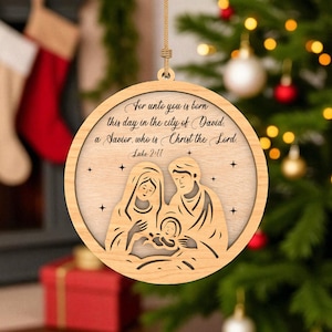 May include: Circular wooden Christmas ornament with a laser-cut Nativity scene. The ornament depicts Mary, Joseph, and baby Jesus, with the text "For unto you is born this day in the city of David, a Savior, who is Christ the Lord. Luke 2:11."