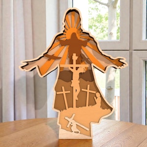 Könnte beinhalten: Ein mehrschichtiges religiöses Kunstwerk aus Holz, das eine zentrale Kreuzigungsszene, eine Figur mit ausgestreckten Armen und mehrere Kreuze darstellt. Das Kunstwerk verwendet warme Töne von Braun, Orange und Beige mit einem silhouettenartigen Design. Es steht auf einem Holztisch.