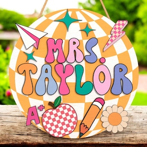 May include: A round wooden sign with the text "MRS. TAYLOR" in colorful bubble letters. The sign has a retro design with a checkered orange and white background, and includes a paper airplane, stars, a lightning bolt, an apple, a pencil, and a flower.