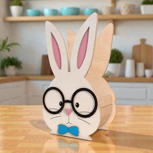 May include: Wooden Easter bunny decoration with a white face, pink inner ears, and a light brown body. The bunny wears black glasses and a blue bow tie. The decoration is on a wooden table.
