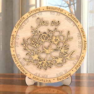 Pode incluir: Placa redonda de madeira com a frase "You are" e um design floral. Palavras inspiradoras e versículos bíblicos circundam o design. Apresentada em um suporte de madeira.