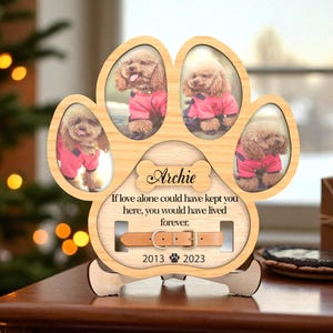 May include: A wooden paw-shaped photo frame with four oval openings, each holding a photo of a dog. The frame includes the name "Archie", the years 2013 and 2023, and a memorial message. A leather collar is also featured.