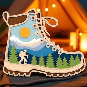 Può includere: Un'opera d'arte in legno a strati a forma di scarpa da trekking. Lo stivale presenta una scena paesaggistica con un escursionista, alberi, montagne e un sole. I colori includono marrone, blu, verde e arancione.