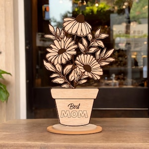 Può includere: Sagoma in legno di una pianta in vaso con fiori e il testo "Best Mom" sul vaso.