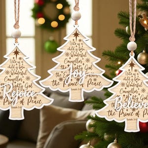 May include: Three wooden Christmas tree ornaments in the shape of a tree, with a light wood grain finish. Each has a white border and features religious text. The ornaments hang from a twine string, perfect for Christmas.